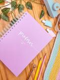 Agenda docente de anillas con portada rosa y la palabra «profesor planner» en blanco, sobre una mesa de madera con lápices