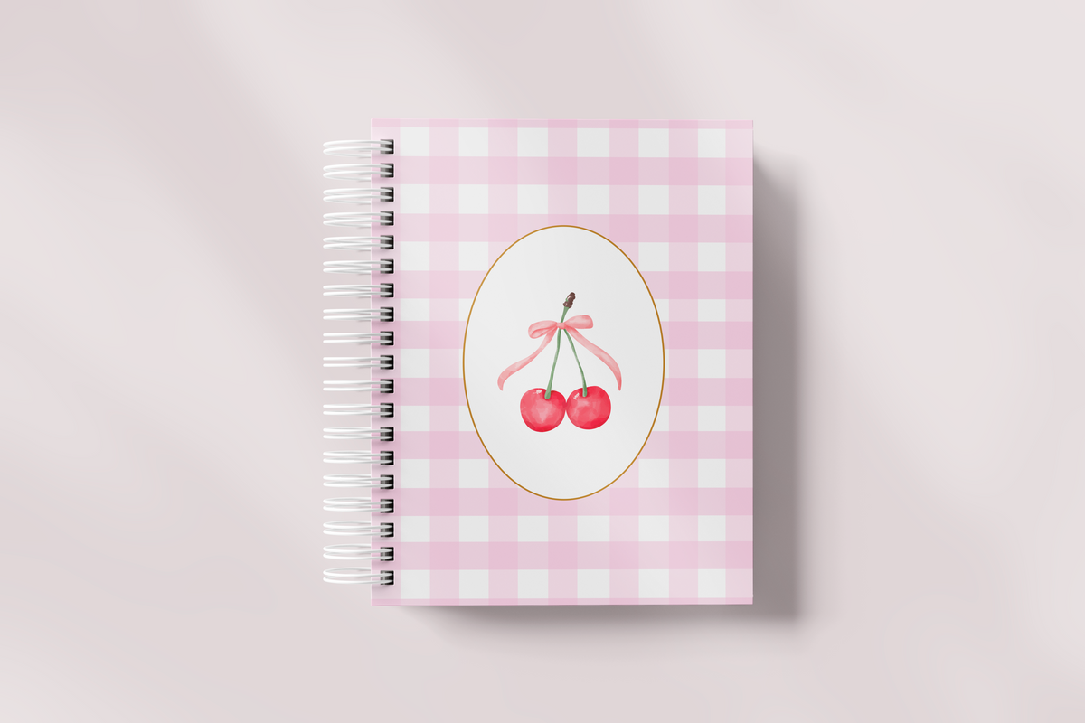 Libreta bonita de anillas blancas con portada de vichy rosa y óvalo con cerezas y lazo, vista de frente sobre fondo rosa claro